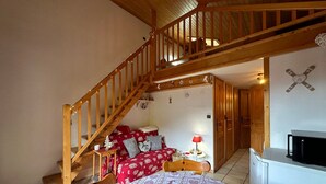 A cozy bedroom with a wooden staircase, a bed with patterned bedding, a nightstand with a lamp, and a TV on a dresser. - Charming duplex for 5 near Blue Lake (Morillon Village)