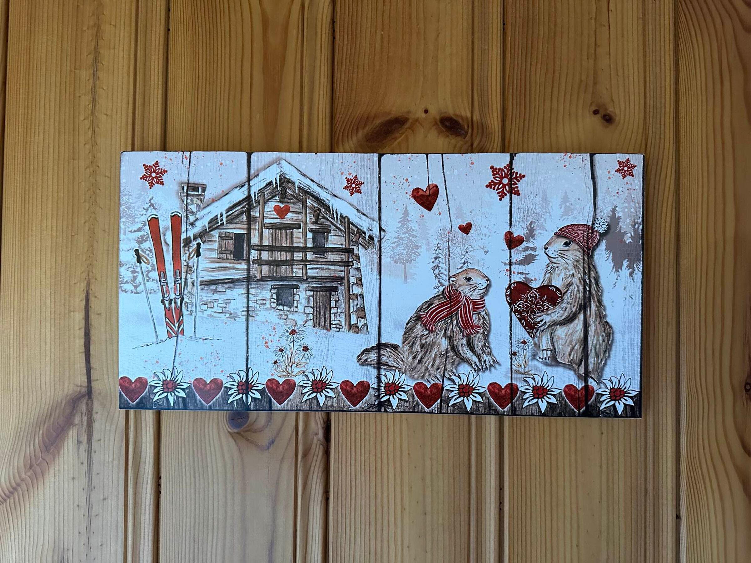 Un panel de madera con una escena invernal, que incluye una cabaña, dos perros, corazones decorativos y flores.