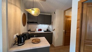 Ett kompakt kök med mörka skåp, en träbänkskiva, en hängande lampa och en dörr som leder till ett annat rum.