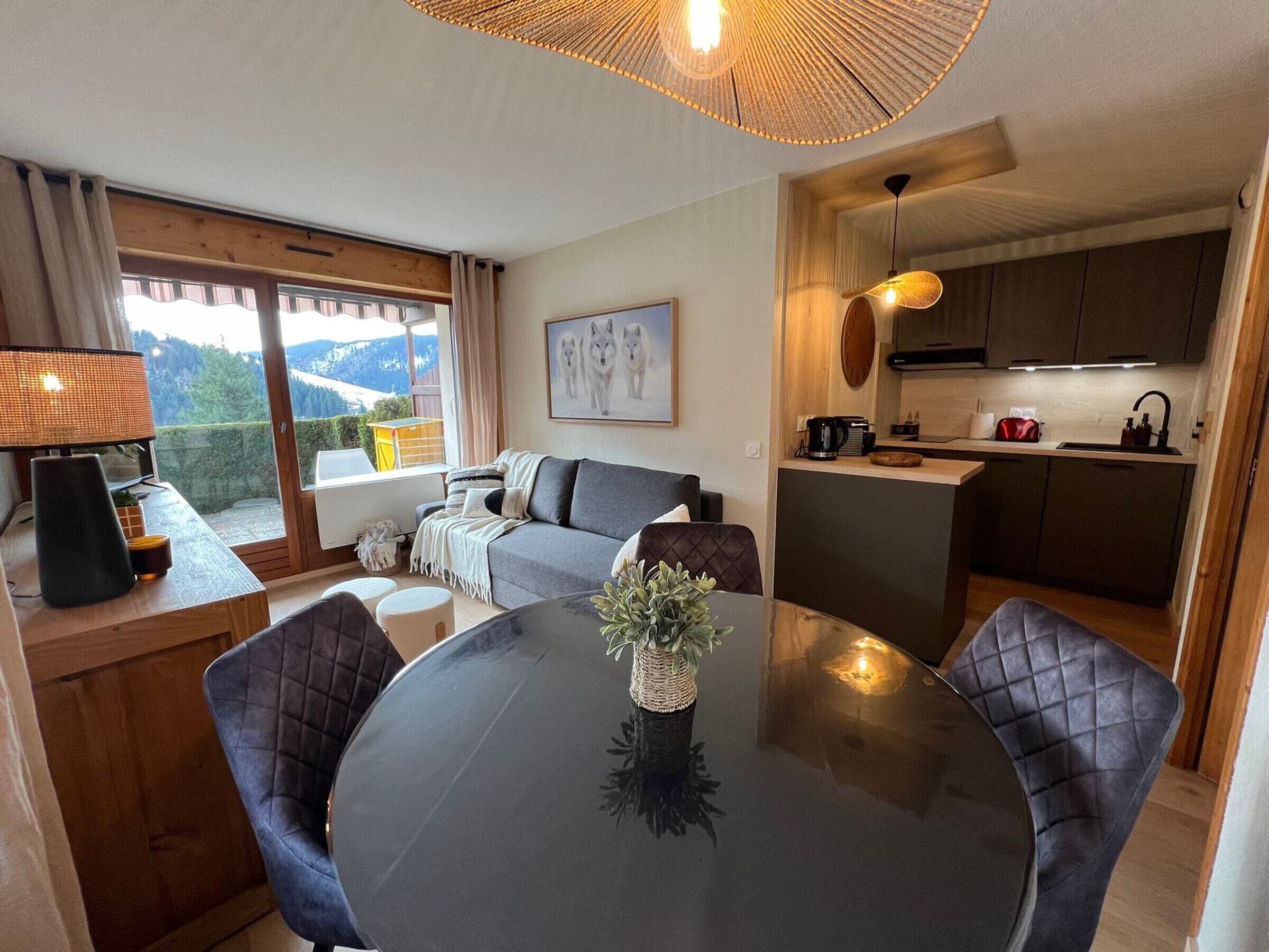 A cozy living area with a sofa, a dining table with chairs, a kitchen with a sink and stove, and a large window with a view of the outdoors.