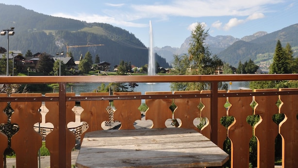 Une terrasse en bois avec une table et des chaises, donnant sur un lac orné d’une fontaine, le tout entouré de montagnes et sous un ciel dégagé.