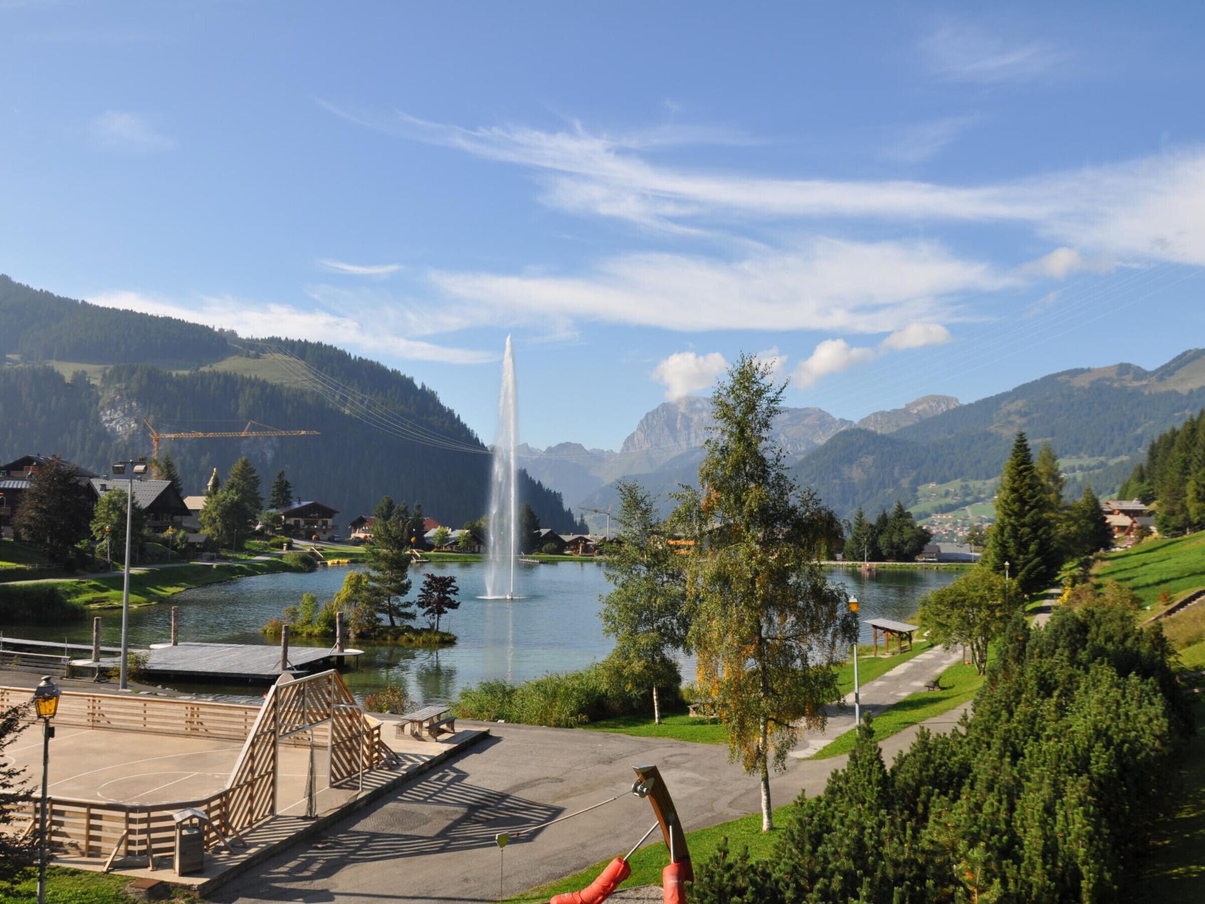 A serene lakeside view with a fountain, a wooden deck, and a mountain backdrop.