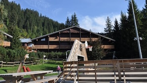 Un chalet en bois avec un toit en pente, entouré d’une forêt. On aperçoit au premier plan un panier de basket et des tables de pique-nique.