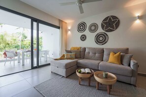 A modern living room with a grey sectional sofa, two wooden side tables, and a glass door leading to an outdoor patio. - Beautiful new beach apartment (Punta Cana)