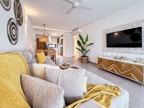 A modern living room with a sofa, a TV, a dining area, and a kitchen in the background. - Beautiful new beach apartment (Punta Cana)