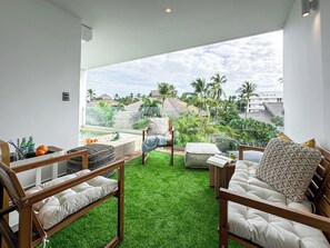 Un salón moderno con suelo de césped artificial verde, mobiliario de madera y un gran ventanal con vistas a palmeras y una piscina.