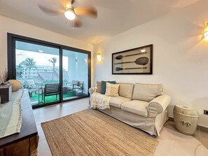 A living room with a sofa, a coffee table, a rug, a ceiling fan, and a sliding glass door leading to an outdoor area with a patio and a pool.
