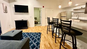 Ett modernt vardagsrum med en platt-TV, en svart möbel för underhållningsutrustning och en grå soffa. Det finns en matplats med ett bord och fyra stolar. Köket har vita skåp och en diskho.