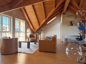 Un loft espacioso con techo alto, vigas de madera, un ventanal grande, un sofá, una mesa de centro y una planta en maceta.