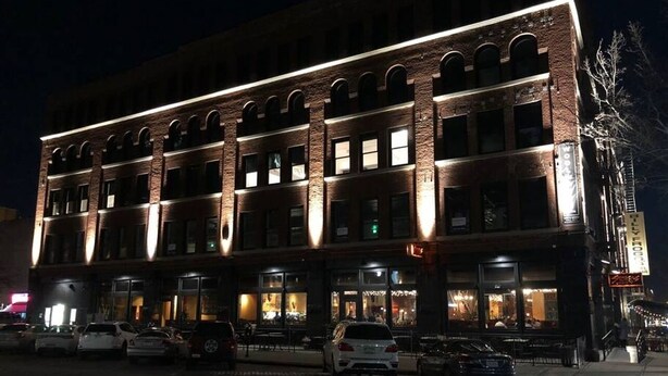 A multi-story building with a brick facade and illuminated windows, featuring a ground-floor restaurant with outdoor seating.