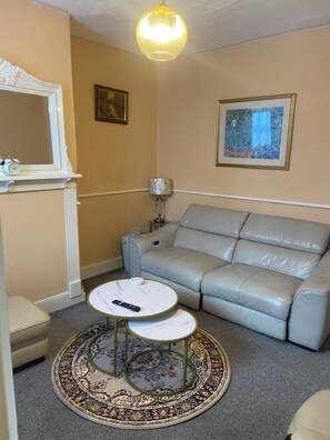 Un salon avec un canapé beige, une table basse ronde, une petite table d’appoint, une lampe, un tableau encadré et un miroir.