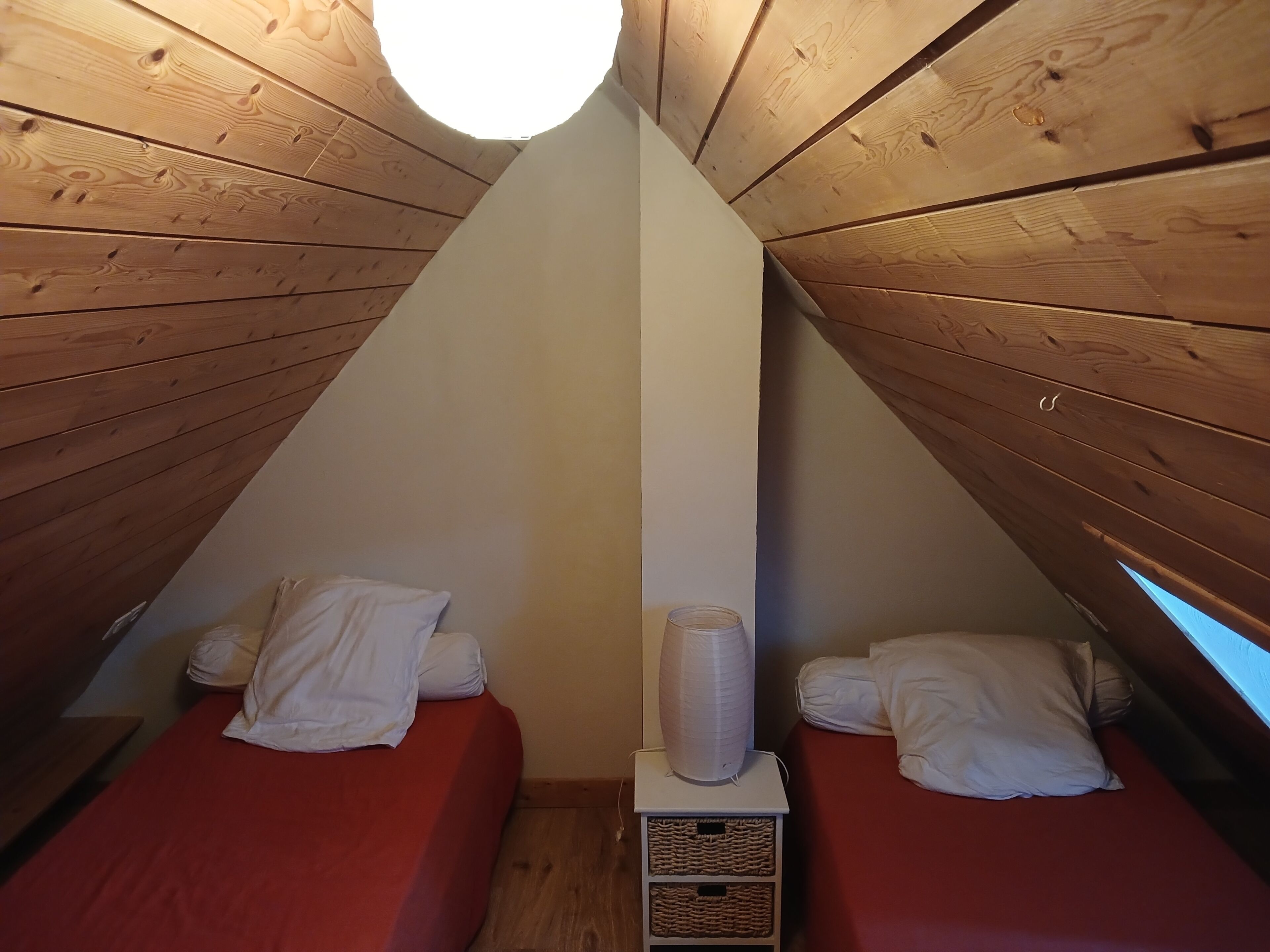 Pequeña habitación de madera con forma triangular, con dos camas, una pared blanca y una lámpara en el techo.
