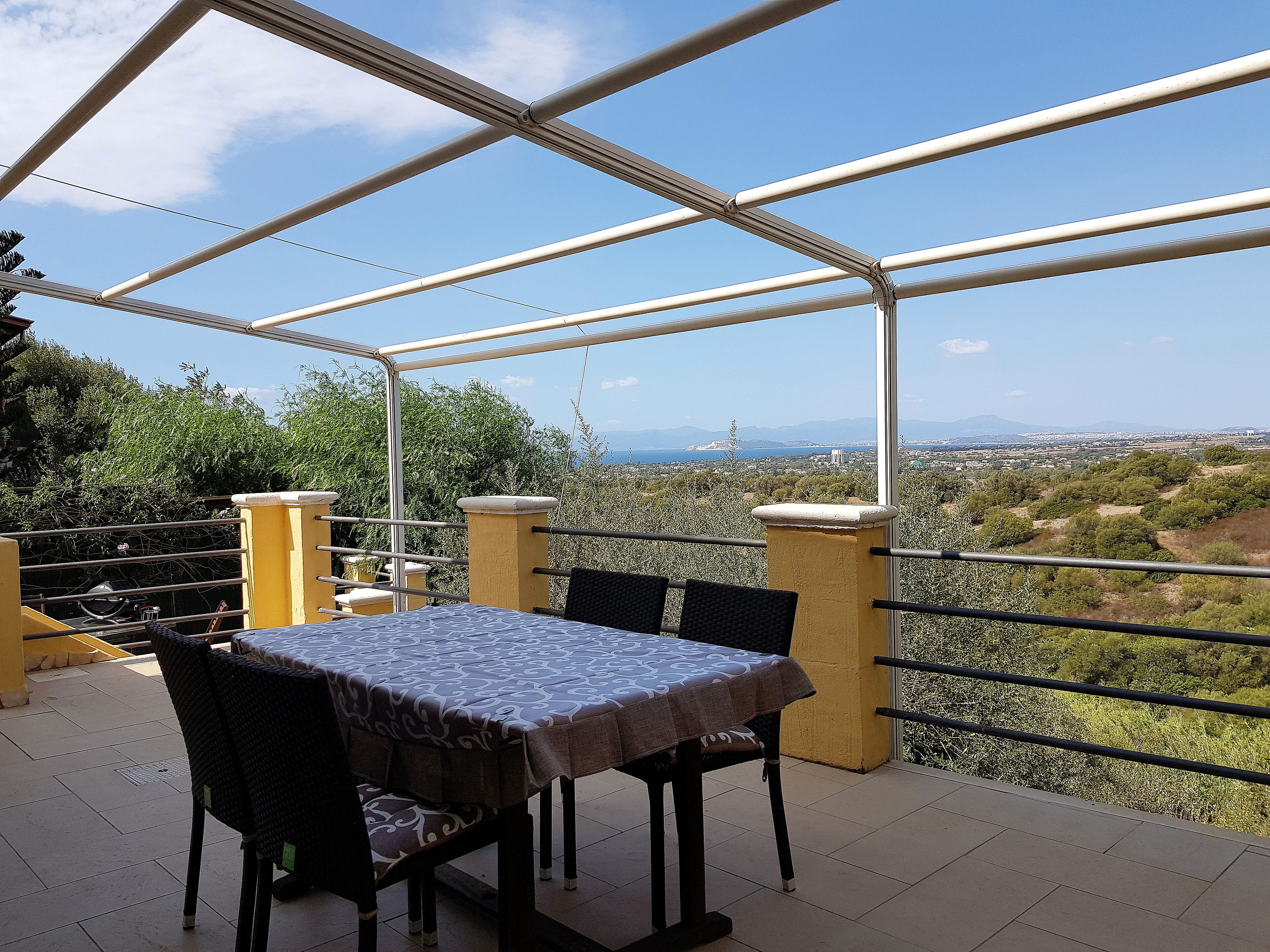An outdoor patio with a table and chairs, a pergola structure, and a view of a landscape with trees and distant mountains.