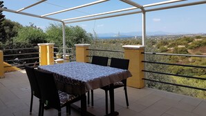 En udendørs terrasse med et bord og stole, en pergola og udsigt til et landskab med træer og fjerne bjerge.