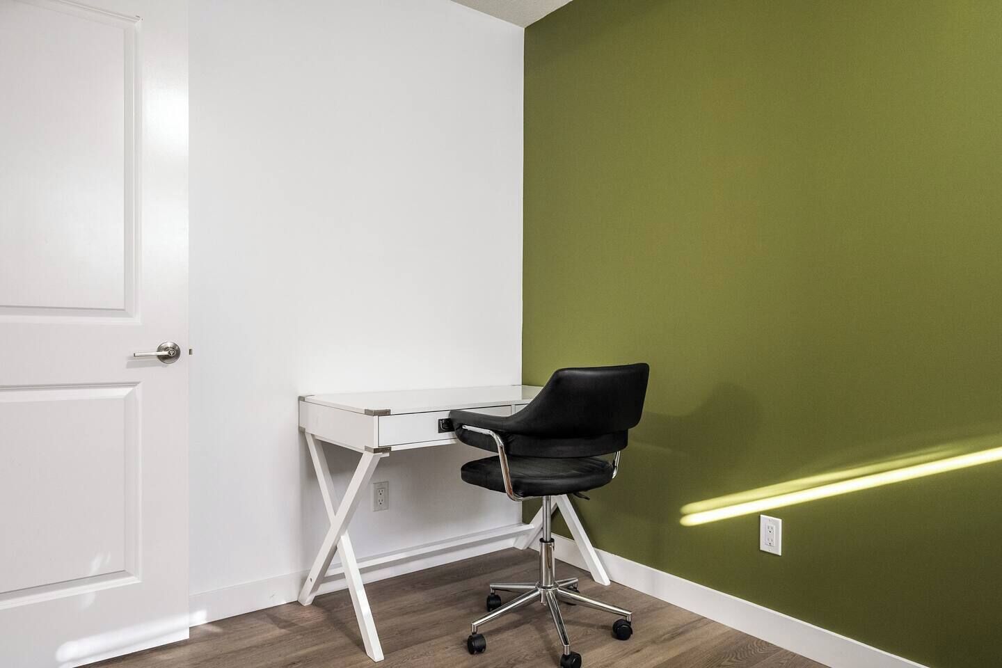 A room with a white desk and black office chair, a green wall, and a white door.