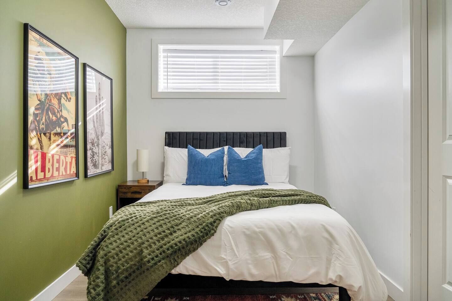 A bedroom with a bed, a nightstand, a lamp, a window with blinds, and framed pictures on the wall.