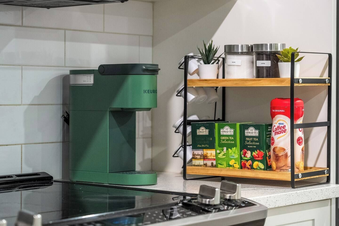 A kitchen with a green Keurig coffee maker, a shelf with Ahmadi tea boxes, and a coffee dispenser.