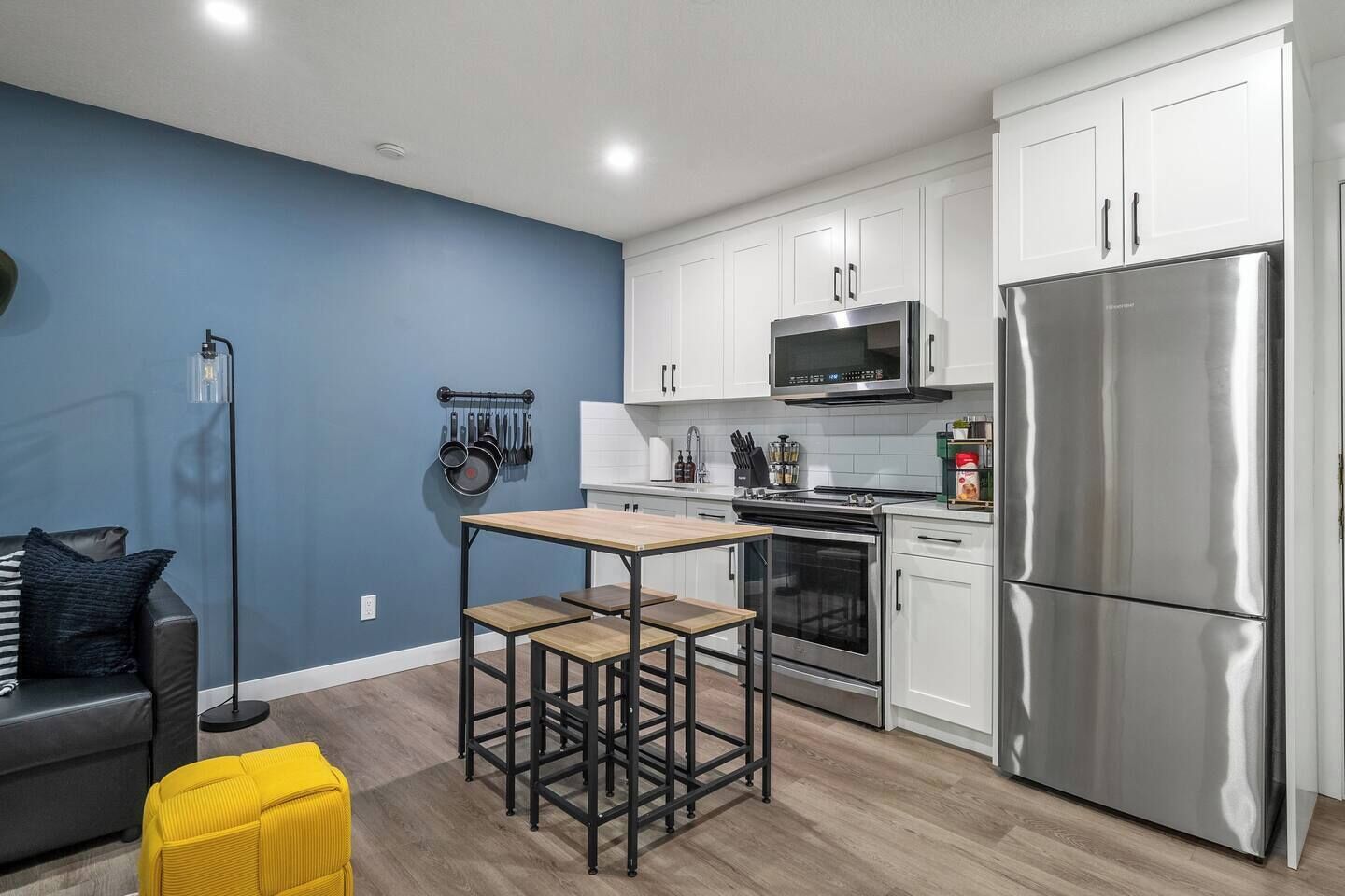 A modern kitchen with white cabinets, a stainless steel refrigerator, and a microwave. There is a dining area with a wooden table and two stools.