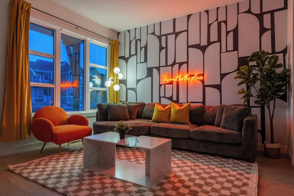 Uma sala de estar com sofá, poltrona, mesa de centro e uma grande janela com cortinas.