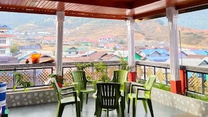 A covered outdoor area with wooden ceiling and walls, green plastic chairs, and a view of a village.