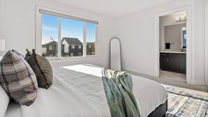 Een slaapkamer met een groot raam, een bed met kussens, een nachtkastje en uitzicht op huizen.