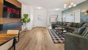 Een moderne woonkamer met een Netflix tv aan een houten wand, een groene bank, een houten salontafel en een geruit tapijt.