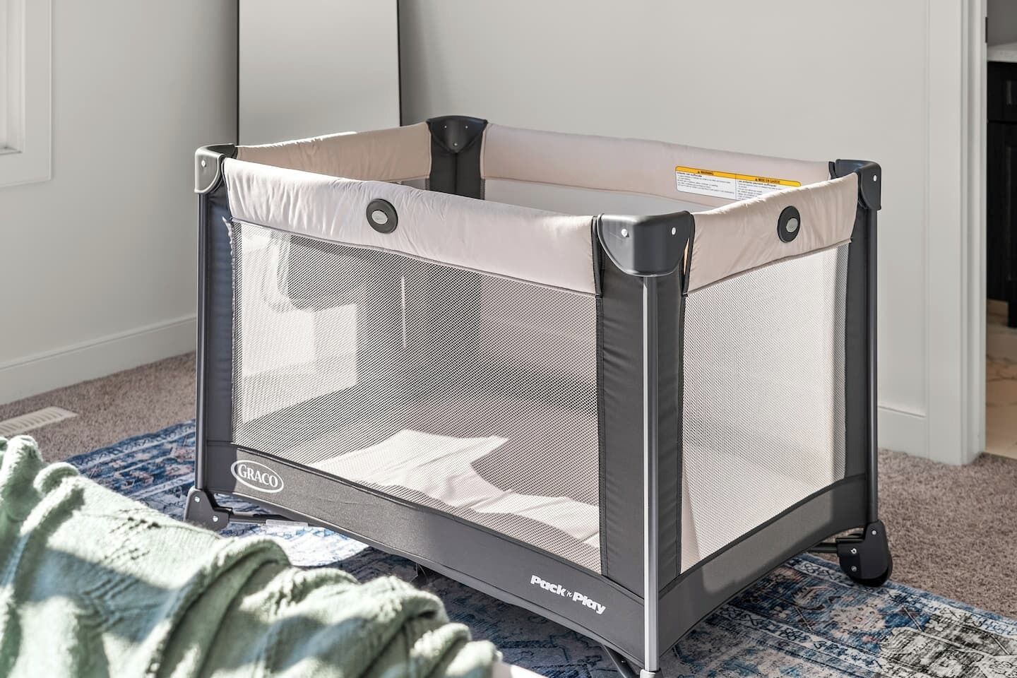 A baby playpen with a mesh side and a padded top, labeled 'GRACO' and 'Puck n Play', placed on a patterned rug.