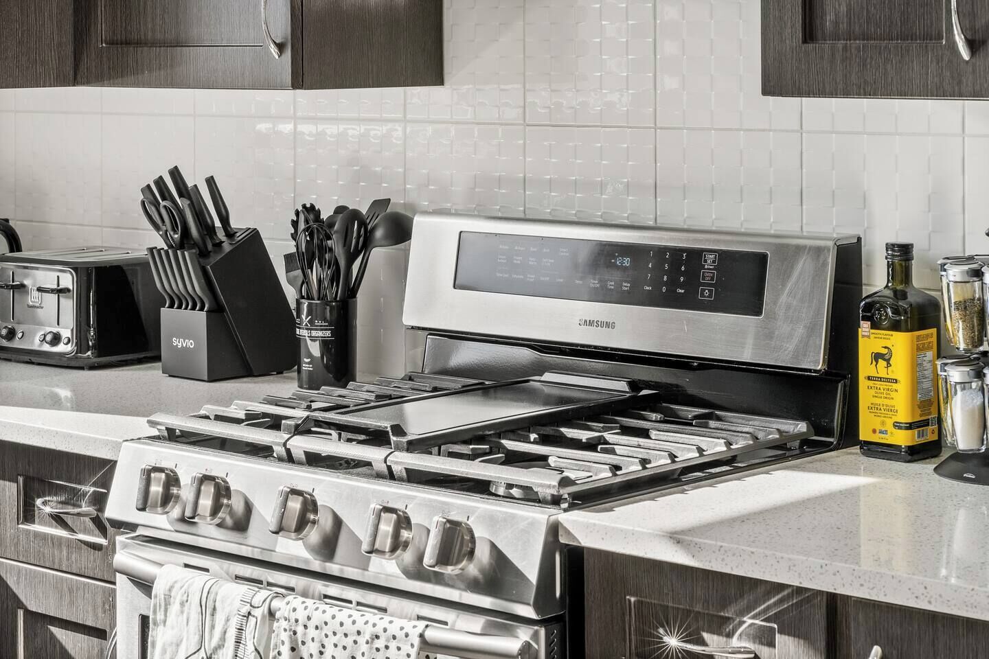 Una cocina con estufa de acero inoxidable, un microondas Samsung y un tostador. Hay utensilios en un organizador y una botella de aceite de oliva sobre la encimera.