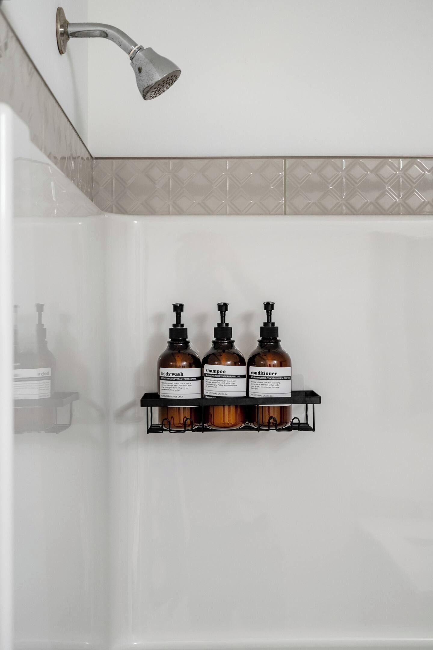 A bathroom with a shower, a white tiled wall, and a shelf holding three bottles of body wash, shampoo, and conditioner.