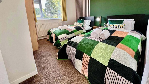 A hotel room with two single beds, a window with curtains, a radiator, and a green wall. - A Vibrant 3-bedroom House With Ample Parking (Derby)