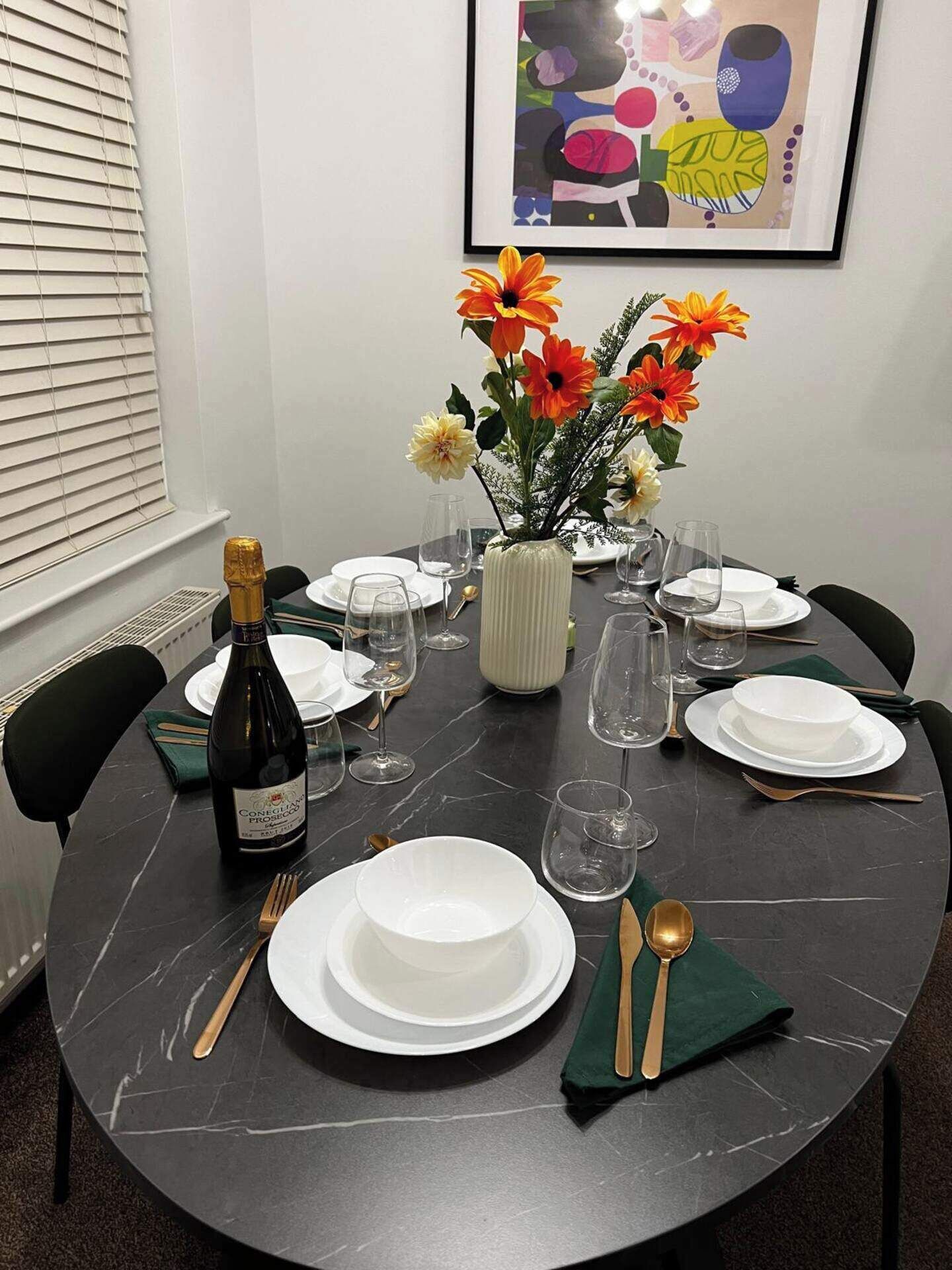 A dining table set with white plates, glasses, and cutlery, a bottle of wine, and a vase with flowers.