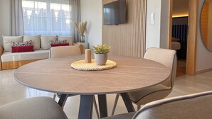 A modern living room with a round dining table, a sofa with patterned cushions, a flat-screen TV mounted on the wall, and a decorative plant.