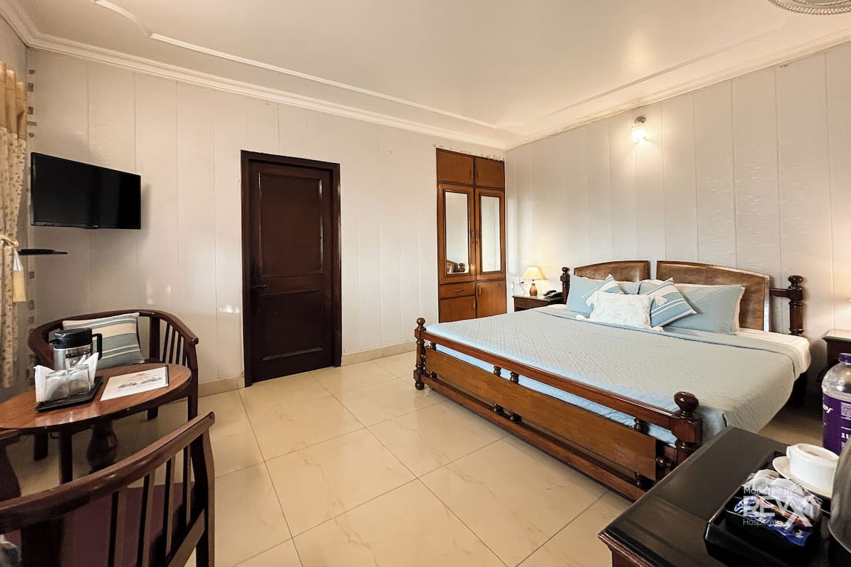 A hotel room with a wooden bed, a small table with a chair, a television mounted on the wall, and a door.