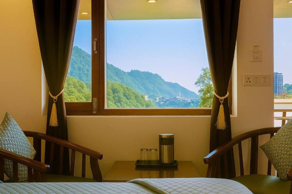 A room with a bed, two chairs, a small table with a kettle, and a window offering a view of green hills.