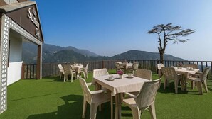 An outdoor dining area with tables and chairs, a view of mountains, and a sign for 'The Sunny Cot'. - Hotel Sunny Cot - Free Mall shuttle and Parking (Dehradun)