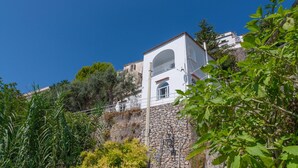 A white building with a stone base, a balcony, and a clear blue sky. - Villa Jacaranda in Praiano (Praiano)
