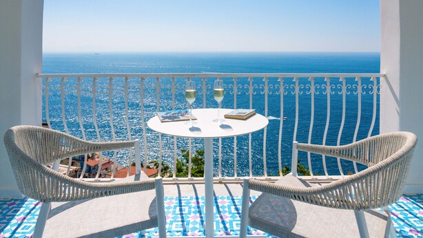 A balcony with wicker chairs, a small round table, and a view of the sea. - Villa Jacaranda in Praiano (Praiano)