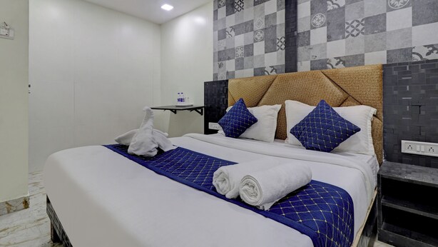 A neatly made bed with white linens and blue accents, a small table with a water bottle, and a patterned wall. - Parle Inn Hotel (Mumbai)