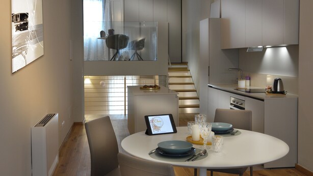 A modern kitchen with a dining area, featuring a tablet on the table displaying a menu, a staircase leading to an upper level, and a view of the outside through a glass door.