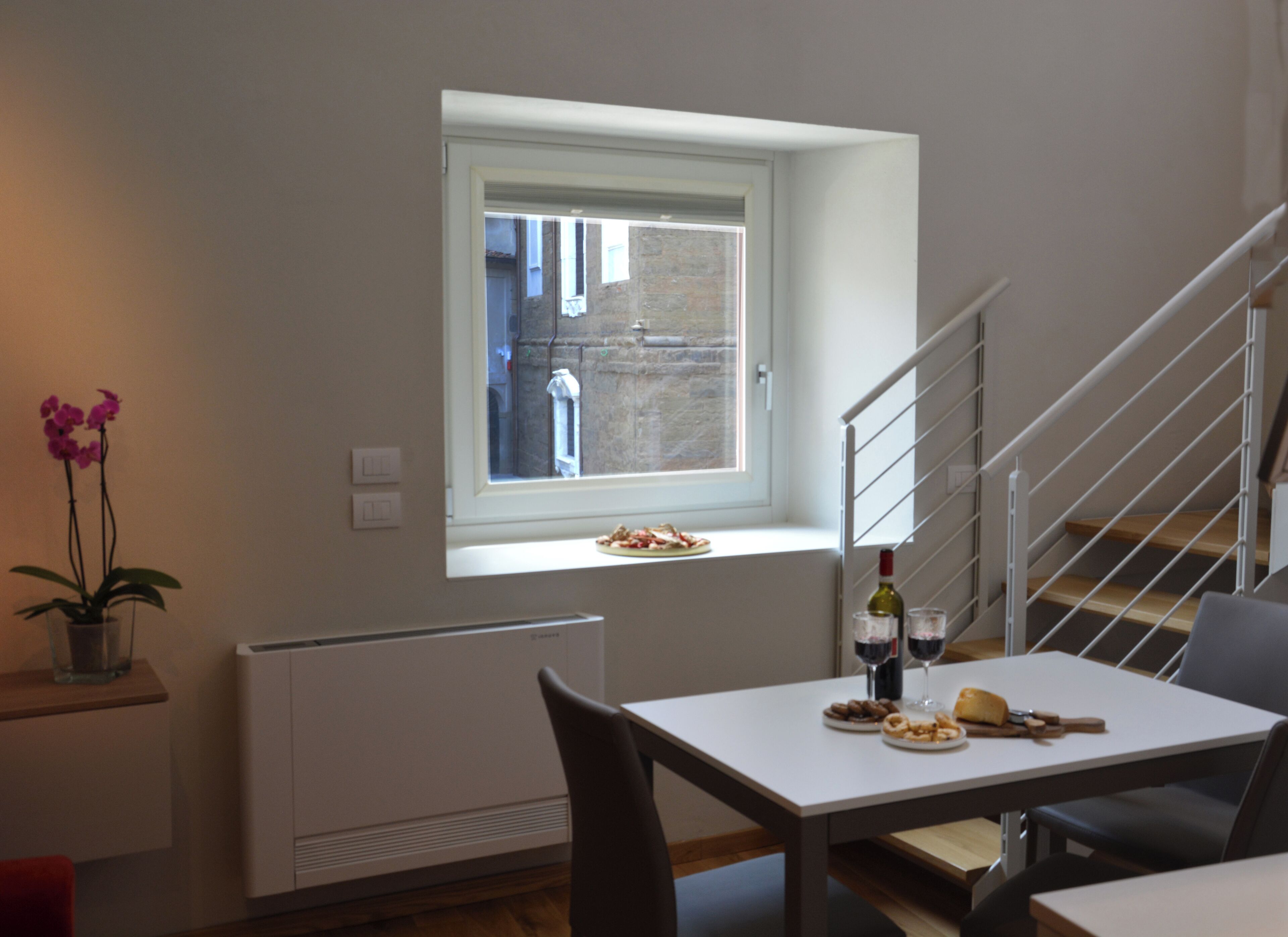 Ein Essbereich mit Tisch für zwei Personen, einem Fenster mit Blick auf ein Backsteingebäude, einer Topfpflanze und einer Heizung.