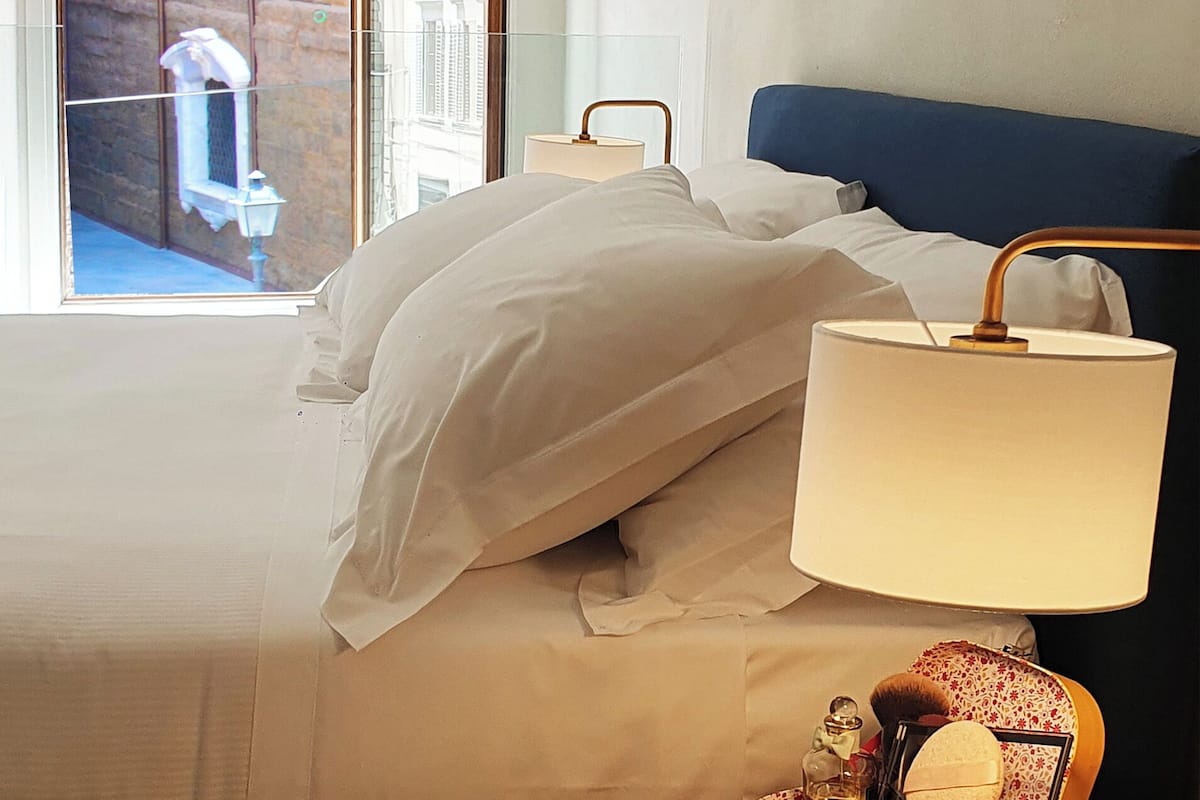A hotel room with a bed, a bedside table with a lamp, a window with a view of the outside, and a painting on the wall.