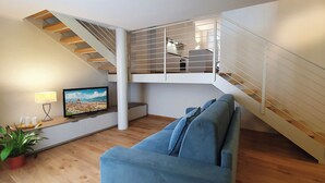 A modern living room with a blue sofa, a TV displaying a cityscape, a staircase with a glass railing, and a potted plant.