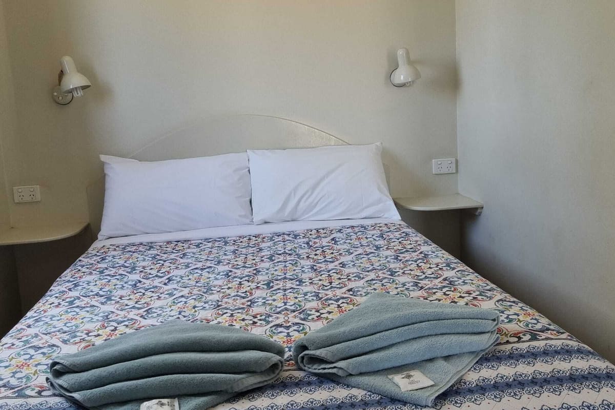 Un lit simple avec une couvre-lit à motifs, deux oreillers et une serviette pliée posée sur le lit. Deux lampes de chevet sont fixées au mur, de part et d’autre du lit.