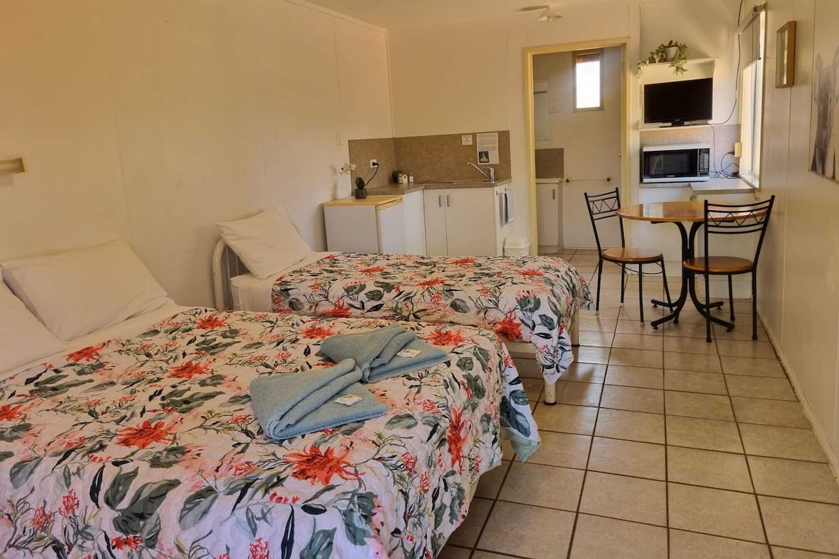 Une chambre d’hôtel compacte comprenant deux lits simples, une petite kitchenette, un coin repas avec une table et des chaises, et un téléviseur fixé au-dessus du comptoir.