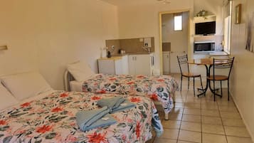 Una habitación de hotel compacta con dos camas individuales, una kitchenette pequeña, una zona de comedor con mesa y sillas, y un televisor montado sobre la encimera.