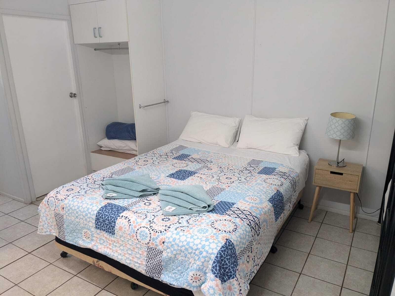 Une chambre à coucher avec un lit, une table de chevet avec une lampe, une armoire et une étagère sur laquelle est posée une serviette bleue.
