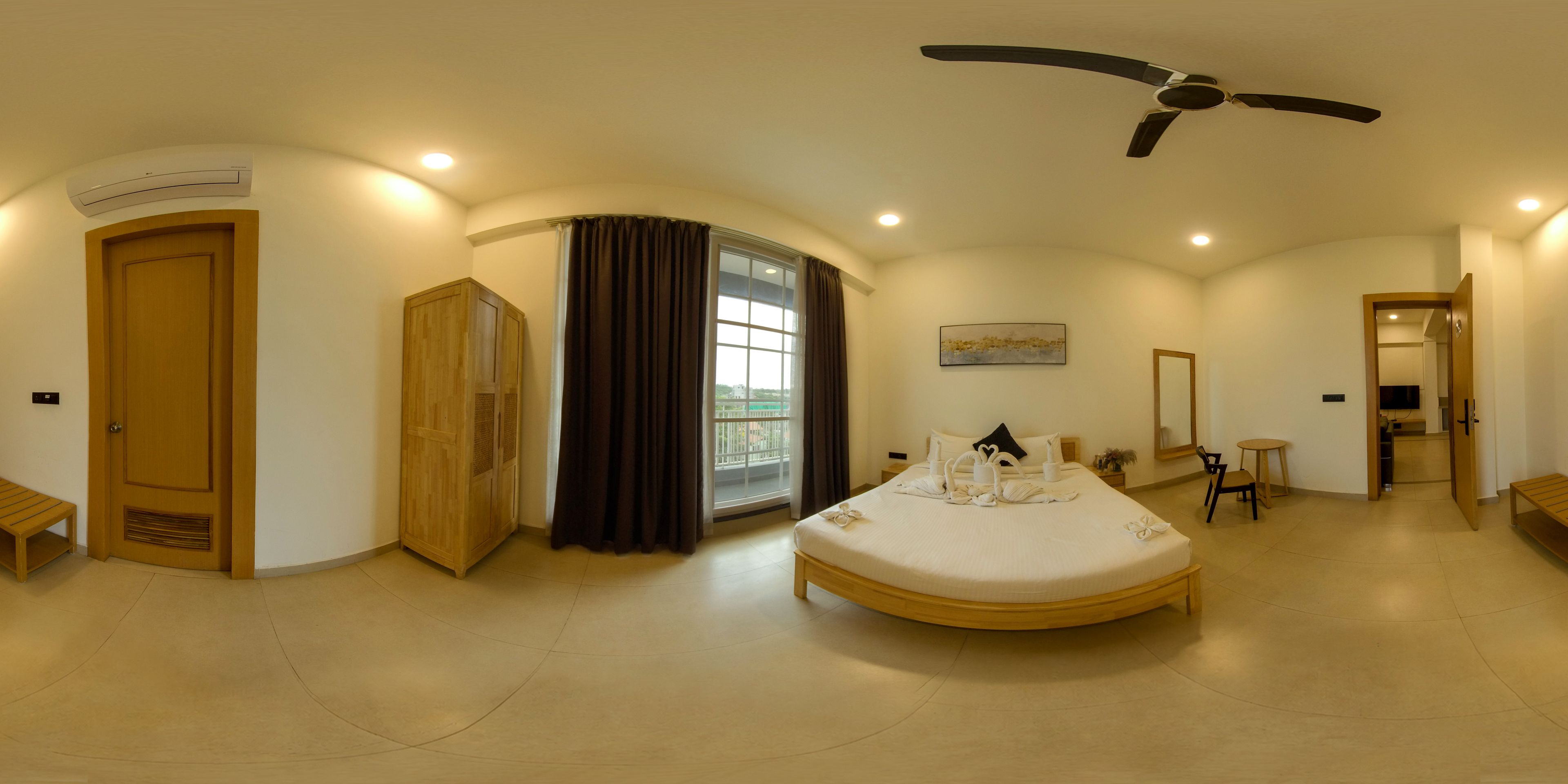Habitación de hotel con cama curvada, ventilador de techo, ventana con cortinas, mesita de noche, espejo y una pequeña mesa con una silla.