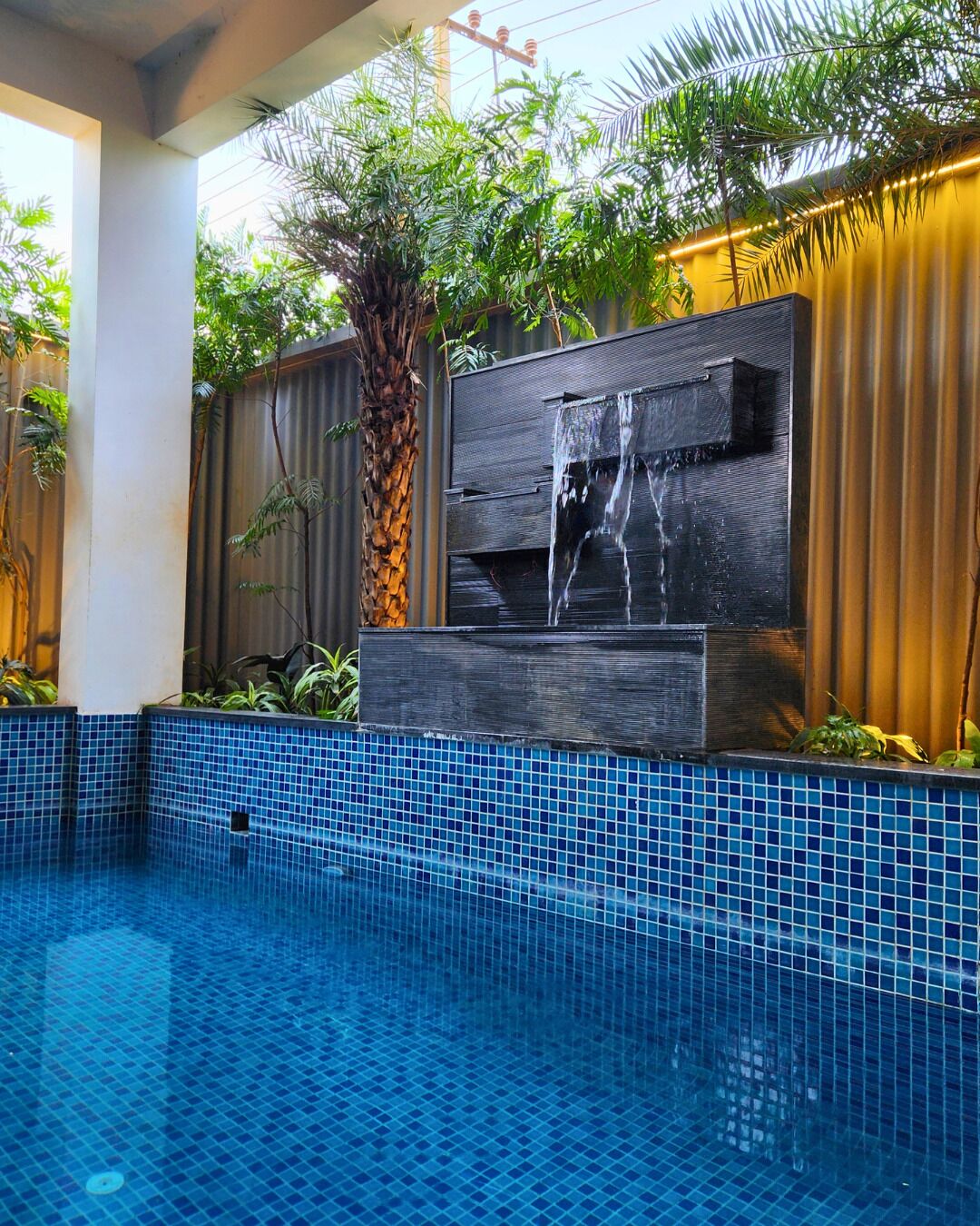 A swimming pool with blue mosaic tiles, a waterfall feature, and tropical plants.