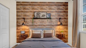 A bedroom with a wooden wall, a bed with two pillows, a bedside table with a clock, and a hanging light fixture.