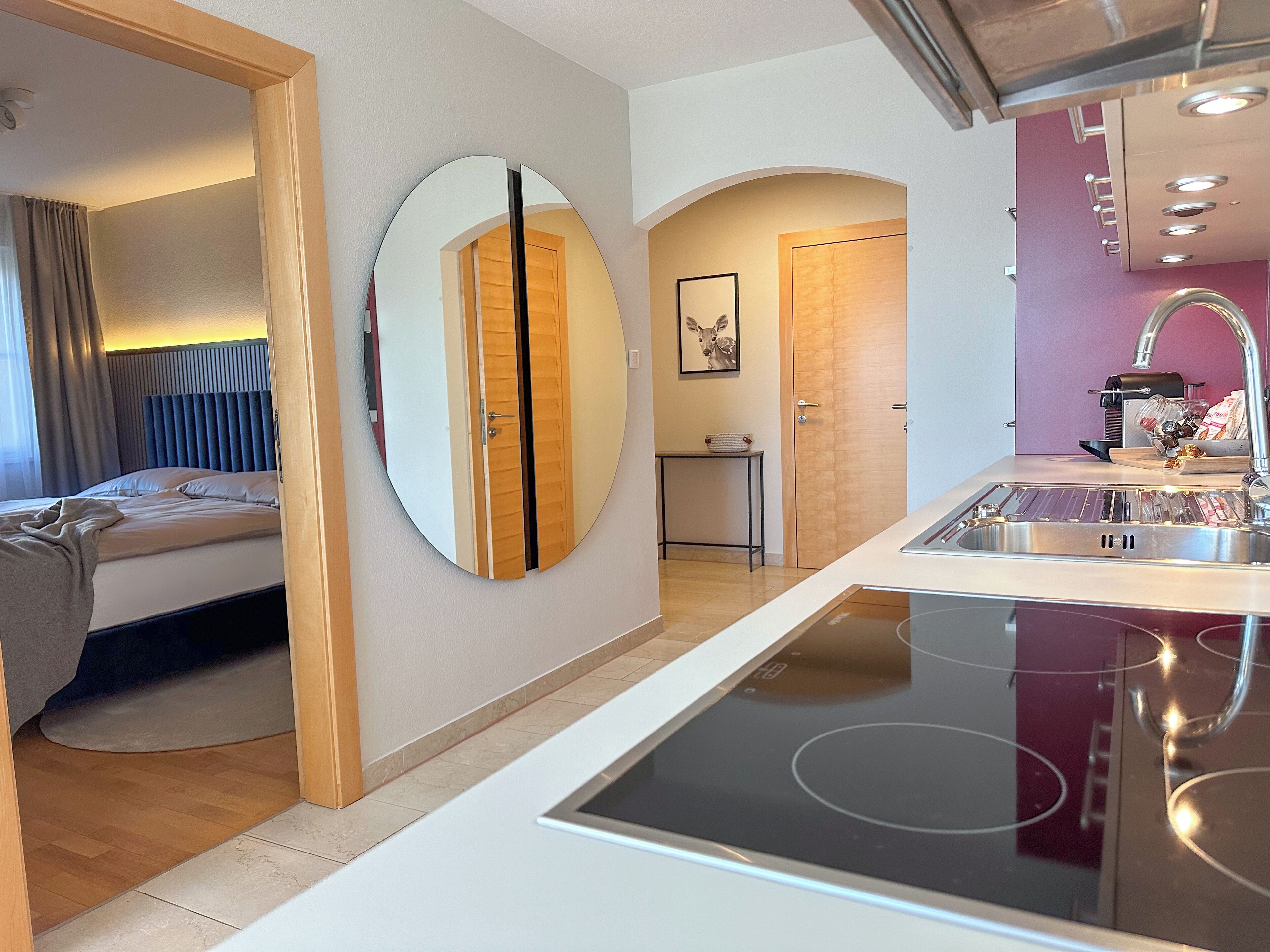 A modern hotel room with a bed, a round mirror, a kitchen with a sink, and a door leading to another room.